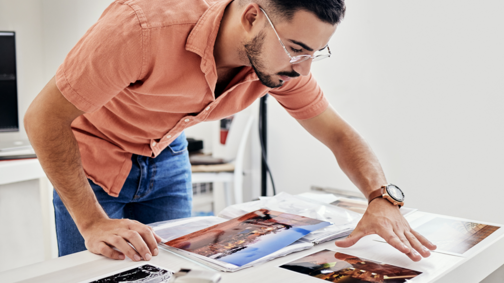 A man in a peach shirt and glasses leans over a table, examining colorful landscape photos.