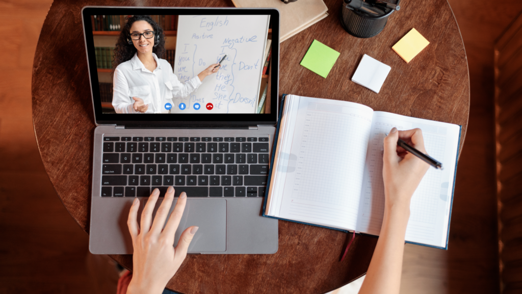 A person watching an online tutor from a laptop and writing down information in a notebook.