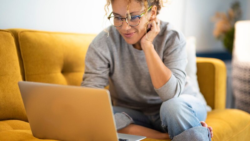 A woman sitting on a yellow couch, browsing on a laptop with a thoughtful expression. She has curly hair and is wearing glasses.