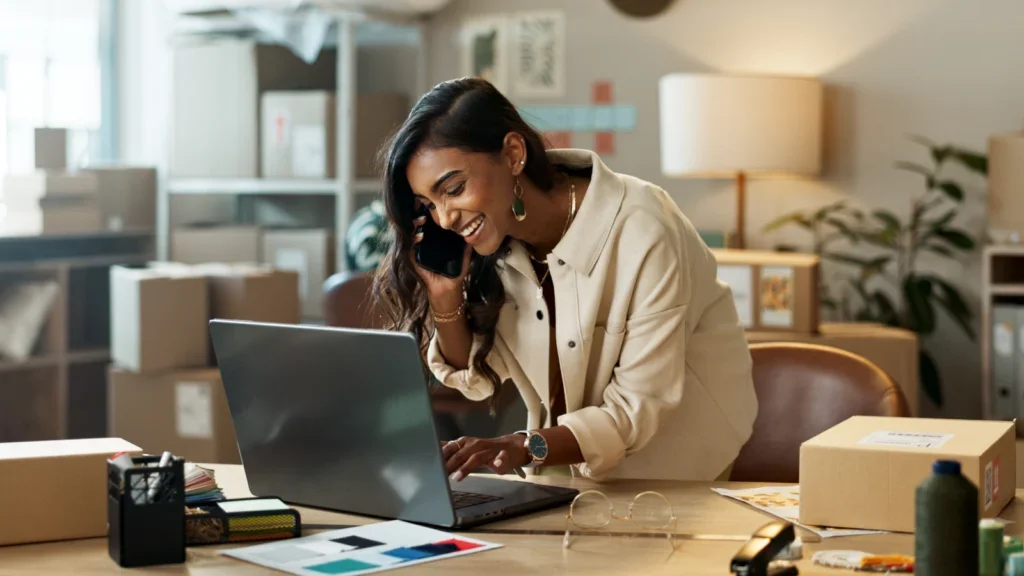 A woman in a beige jacket smiles while talking on the phone and typing on a laptop in a home office. Cardboard boxes and greenery around her.
