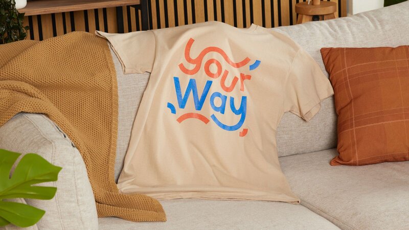 A camel t-shirt with blue and red “Your way” text lying on top of a sofa.