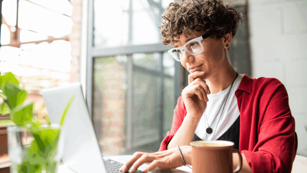 A woman in glasses with curly hair is sitting at a table with a laptop and reading about hobbies that make money.