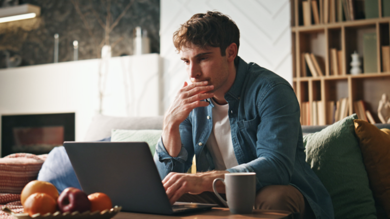 A man in a denim shirt is sitting on a sofa, focused on a laptop in front of him, searching for advanced TikTok affiliate marketing strategies.