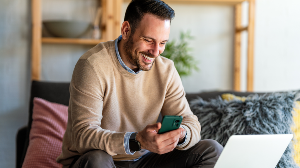A smiling man sitting on a couch in front of a laptop with a mobile phone in his hands.