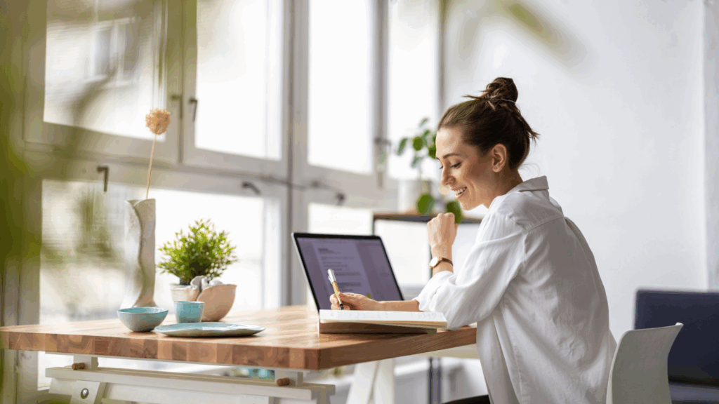 A smiling woman in a white blouse sitting at a desk with a laptop on top of it, making notes.