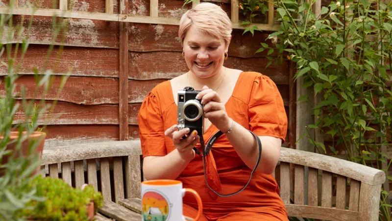 A smiling woman in an orange dress is taking a picture of a colorful mug using a camera in a garden.