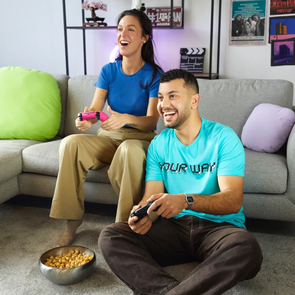Two friends playing video games together, having fun and eating popcorn, wearing print on demand t-shirts.