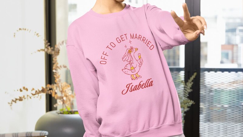 A woman wearing a pink sweatshirt with the text "OFF TO GET MARRIED" and a graphic of a duck, along with the name "Isabella."