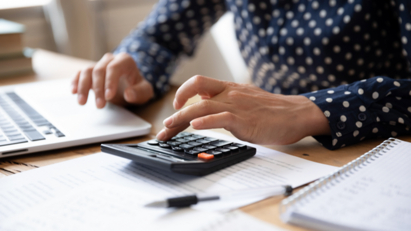 Close-up of hands using a calculator and a laptop on a desk with papers and notebooks. The person wears a polka dot shirt.