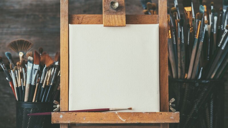 Blank canvas on a wooden easel surrounded by cups of paintbrushes.