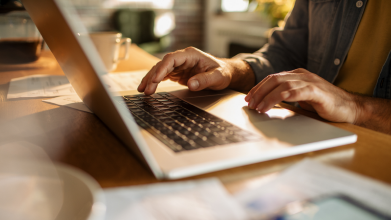 Hands typing on a laptop at a wooden desk, surrounded by papers and a coffee cup.