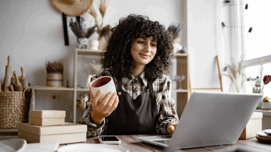 A woman with curly hair sits at a table, focused on her laptop, ready to sell her art online.