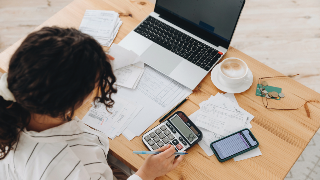 A woman using a calculator while sitting at a desk full of different papers and a laptop.