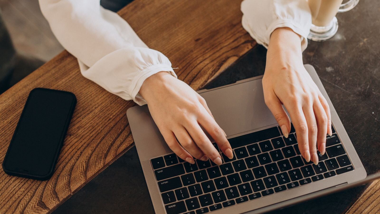 A person's hands typing on a laptop keyboard, positioned on a wooden table. A smartphone is placed beside the laptop.