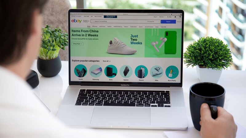 A person sitting at a desk, looking at a laptop displaying the eBay homepage with various products, while holding a black mug.