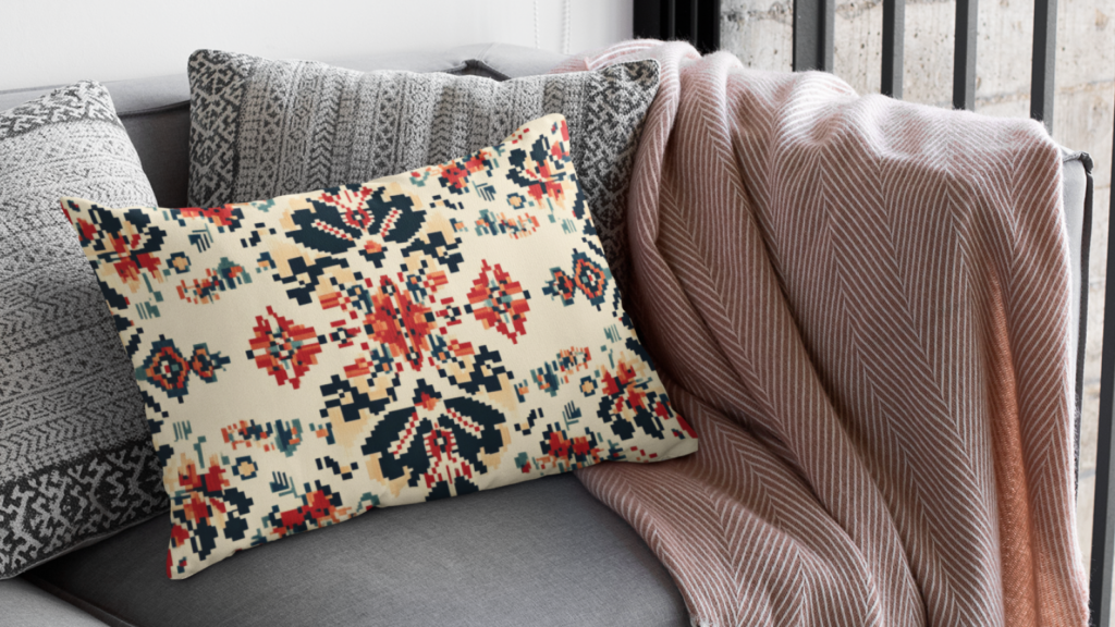A patterned pillow with red, navy, and cream designs on a grey couch, alongside a textured grey pillow and a draped blanket.