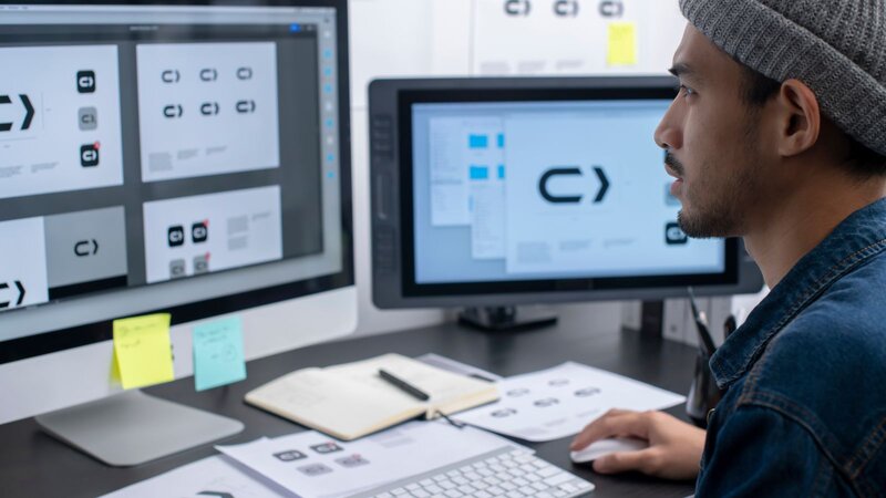 A man wearing a beanie and a denim jacket sits at a desk, working on graphic design software, with multiple screens.