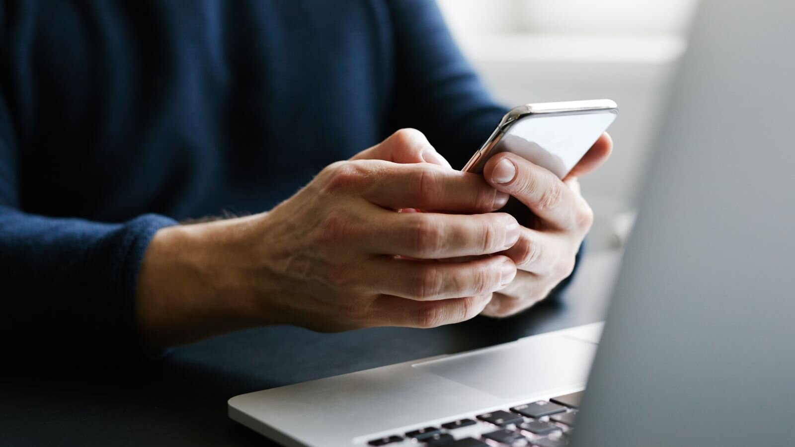 A person in a dark sweater uses a smartphone, focusing intently while sitting at a desk with a laptop.