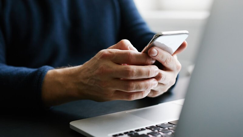 A person in a dark sweater uses a smartphone, focusing intently while sitting at a desk with a laptop.