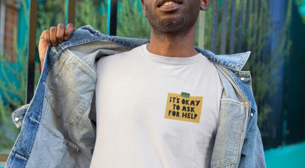 A man in a t-shirt displaying the phrase "it's okay to ask for help" stands confidently.