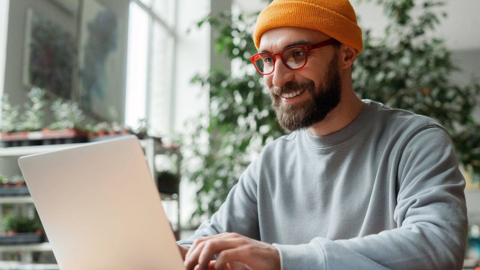 A smiling man with a beard, wearing a gray sweatshirt and orange beanie, working on a laptop, exploring Wix eCommerce features.