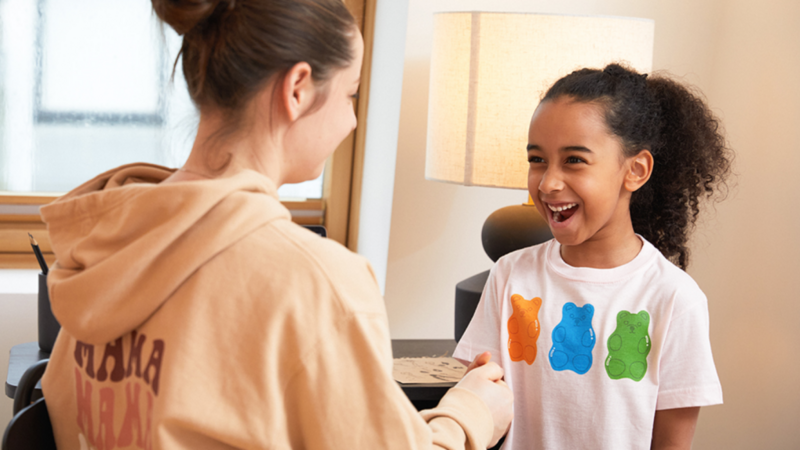A woman in a tan hoodie smiles at a laughing girl wearing a white shirt with colorful bear designs.