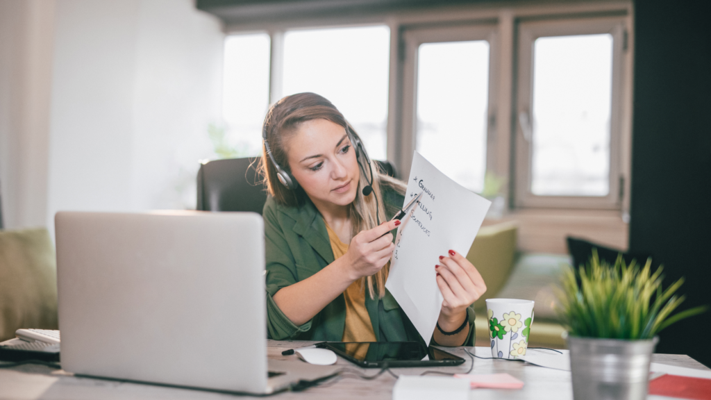 A woman is seated at a desk, using her laptop, with a piece of paper beside her, indicating a work environment.