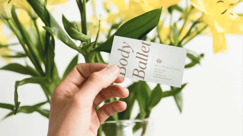 A woman is holding a “Body Balley” business card with yellow flowers in the background.