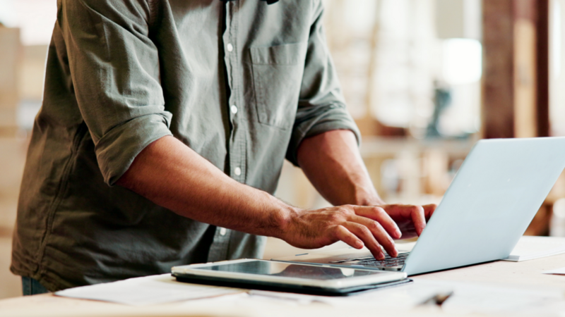 A close-up shot of a man’s hands typing on a laptop. A tablet lies next to the laptop.