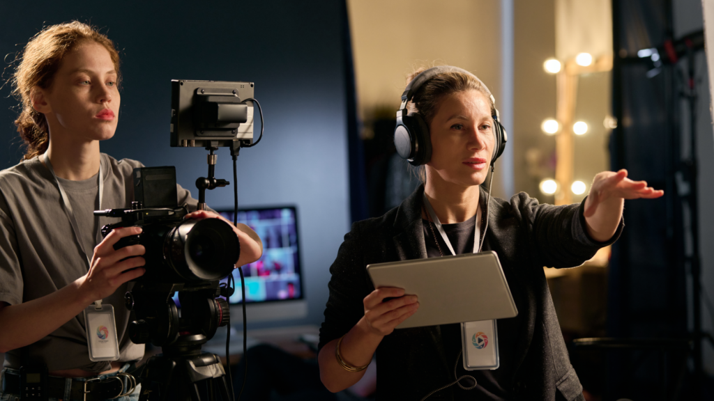 Two women stand in front of a camera and a laptop, capturing a moment of teamwork and creativity.