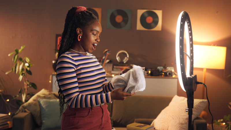 A surprised woman in a striped long-sleeve shirt is unwrapping a product and recording it using a smartphone with a ring light.
