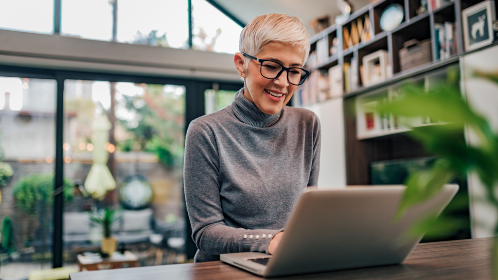 A woman wearing glasses is focused on using a laptop at a desk, with a thoughtful expression on her face.