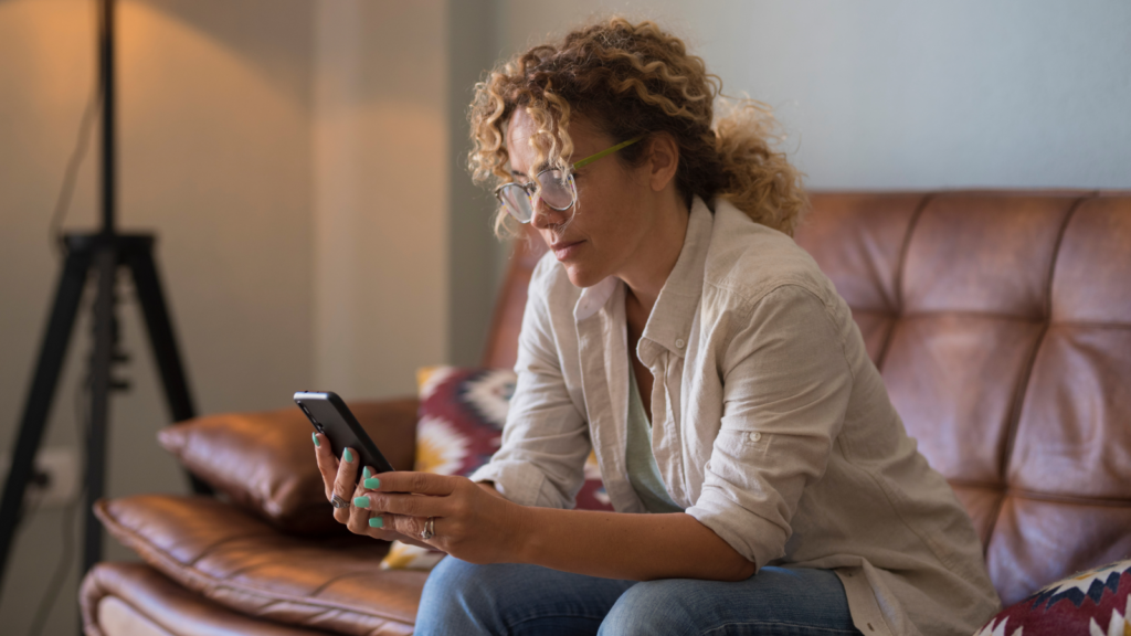 A woman in glasses sitting on a leather couch and looking at her mobile phone.