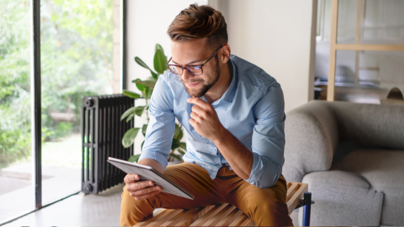 A man in glasses and a blue shirt sits on a coffee table, looking at a tablet. A couch and a large window with greenery are in the background.