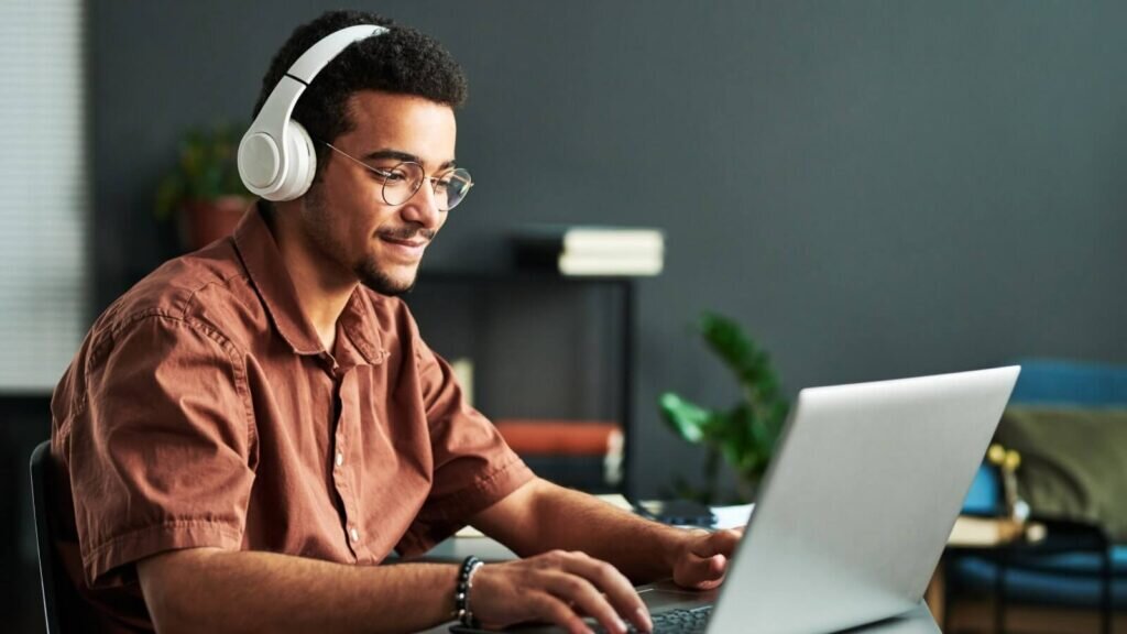 A man sitting at a desk and looking at a laptop screen, wearing headphones.