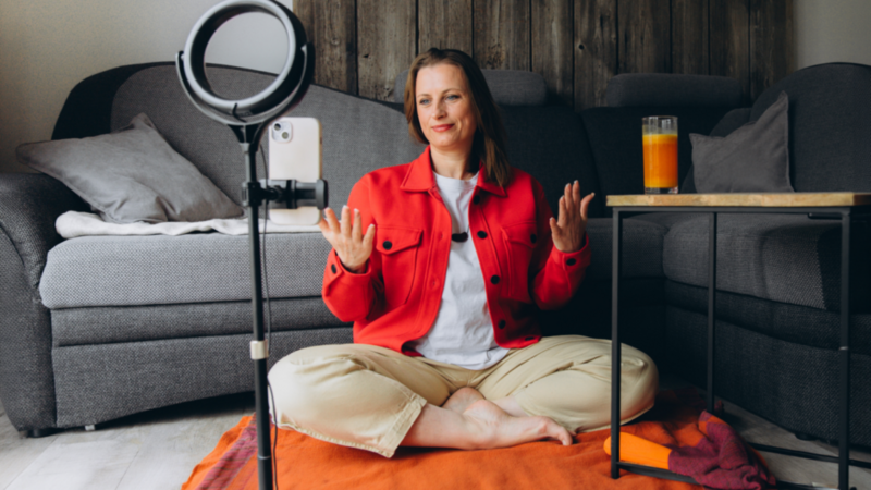 A woman in a red jacket sits cross-legged on an orange mat in a cozy living room, gesturing while filming with a smartphone on a tripod.
