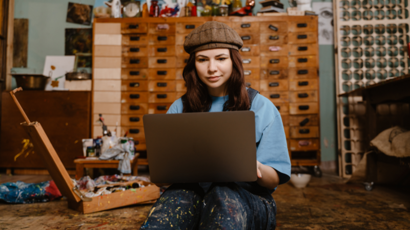 A woman in a blue shirt and beret sits on the floor of an art studio, focused on a laptop, searching for how much you can make on Etsy.