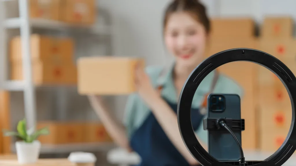A mobile phone on a stand in front of a blurred woman holding a box and filming herself.