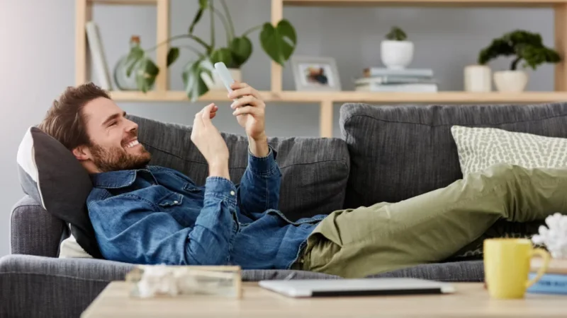 A man in a denim shirt and khaki pants lounges on a gray sofa, smiling at his smartphone.
