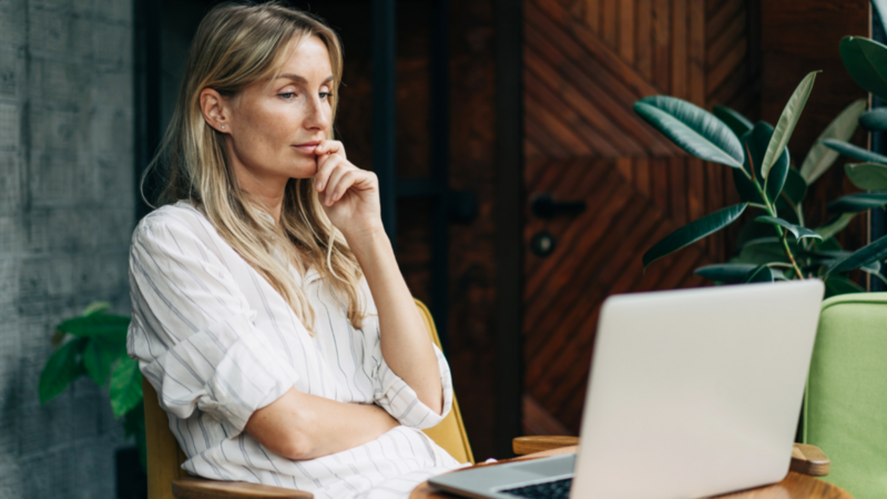 A woman with long blonde hair, wearing a striped shirt, looks at a laptop. Seated indoors with a potted plant and wooden decor.