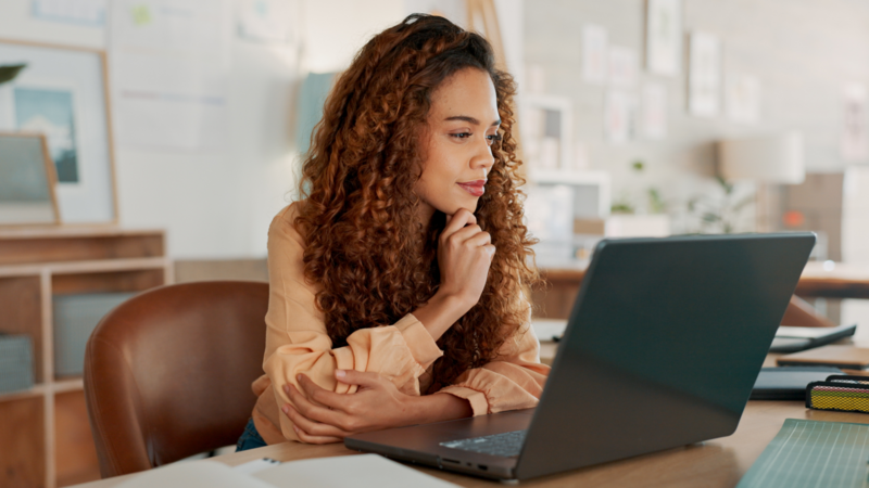 A young woman with curly hair sitting at a desk, thoughtfully looking at a laptop, reading about how to use Shopify Canvas.