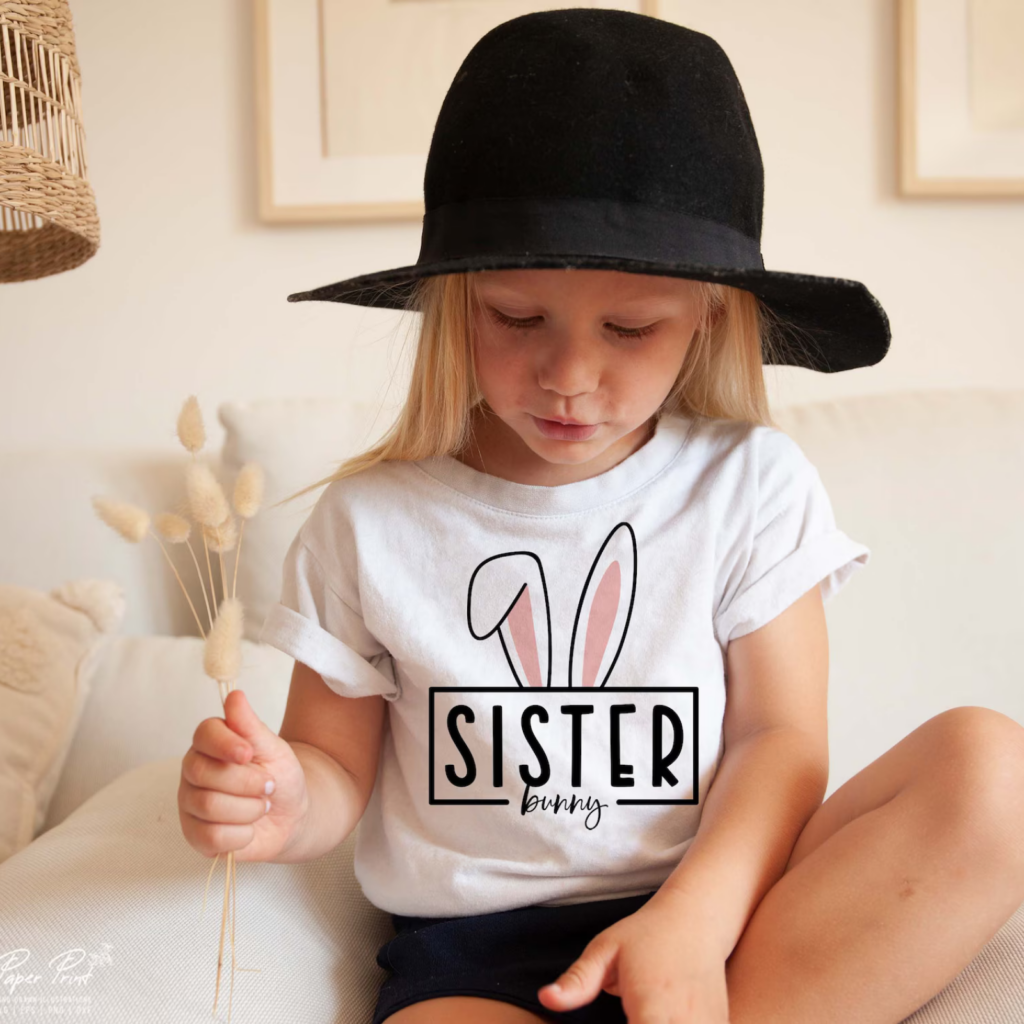 Young girl wearing a floppy black hat and a white shirt with bunny ears and "Sister Bunny" text, holding dried flowers, sitting on a white couch.
