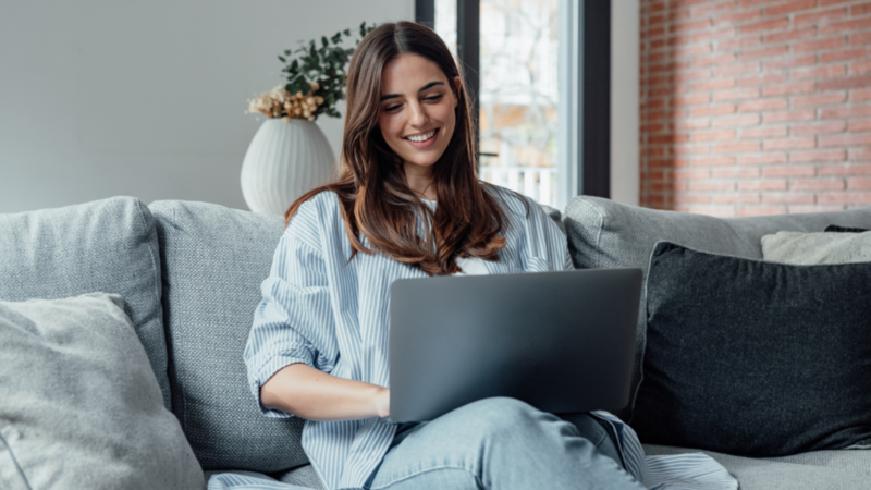 A woman sitting comfortably on a couch, focused on her laptop, with a cozy living room setting in the background.
