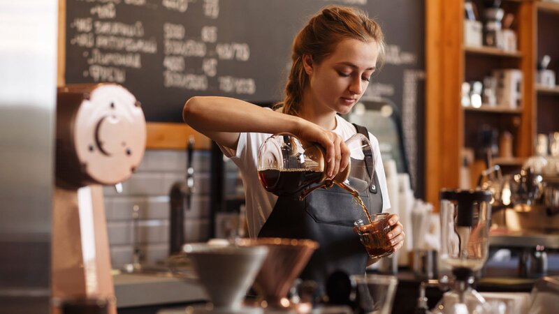 A barista in a gray apron pours pour-over coffee from a glass carafe into a small cup at a café counter with a chalkboard menu behind her.