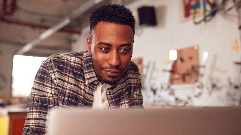 A young man with short hair, wearing a plaid shirt, looking at a laptop screen, learning how to use TikTok for eCommerce.