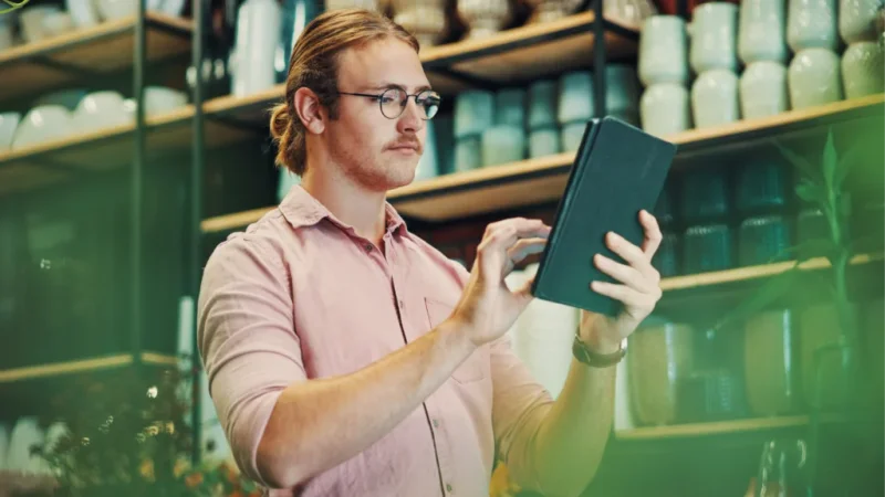 A man wearing glasses is holding a tablet computer, focused on the screen with a thoughtful expression.