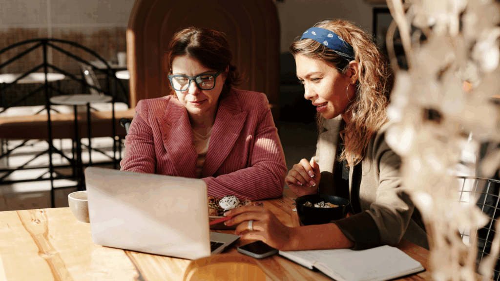 Two women seated at a table, collaborating on a laptop, engaged in discussion and sharing ideas.
