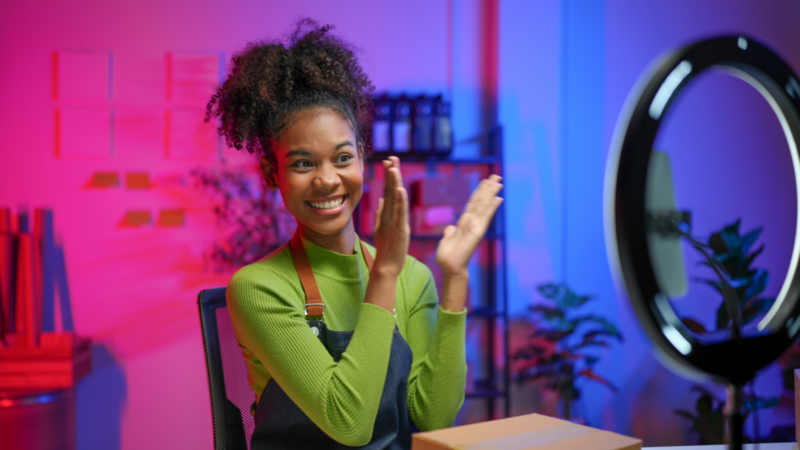 A smiling woman in a green sweater and apron claps happily while live-streaming in a colorful room with pink and blue lighting and plants.