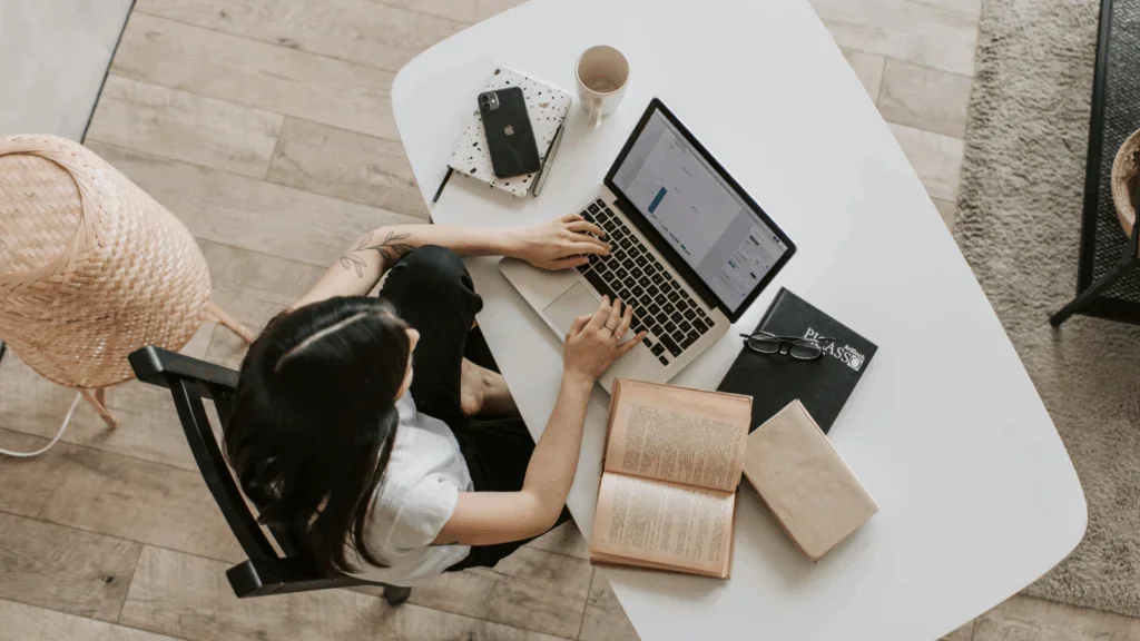 An upper shot of a woman reading and using a laptop at a desk.