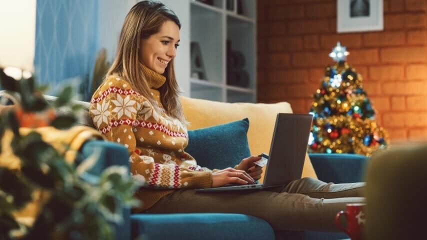 A woman wearing a Christmas sweater and sitting next to a Christmas tree while working on her laptop.